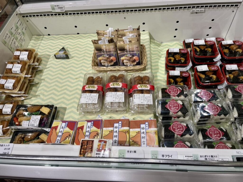 佐野SA上り売店の弁当棚アップ。天むす、おにぎり、豚焼肉弁当など子連れでもシェアしやすい惣菜と弁当が並んでいる